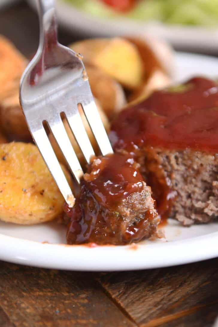 A mini glazed meatloaf with a bite taken out and a fork taking the bite. Roasted potatoes are next to the meatloaf.