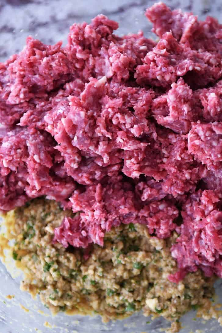 Top down view of raw meat and a bread crumb mixture in a glass bowl.