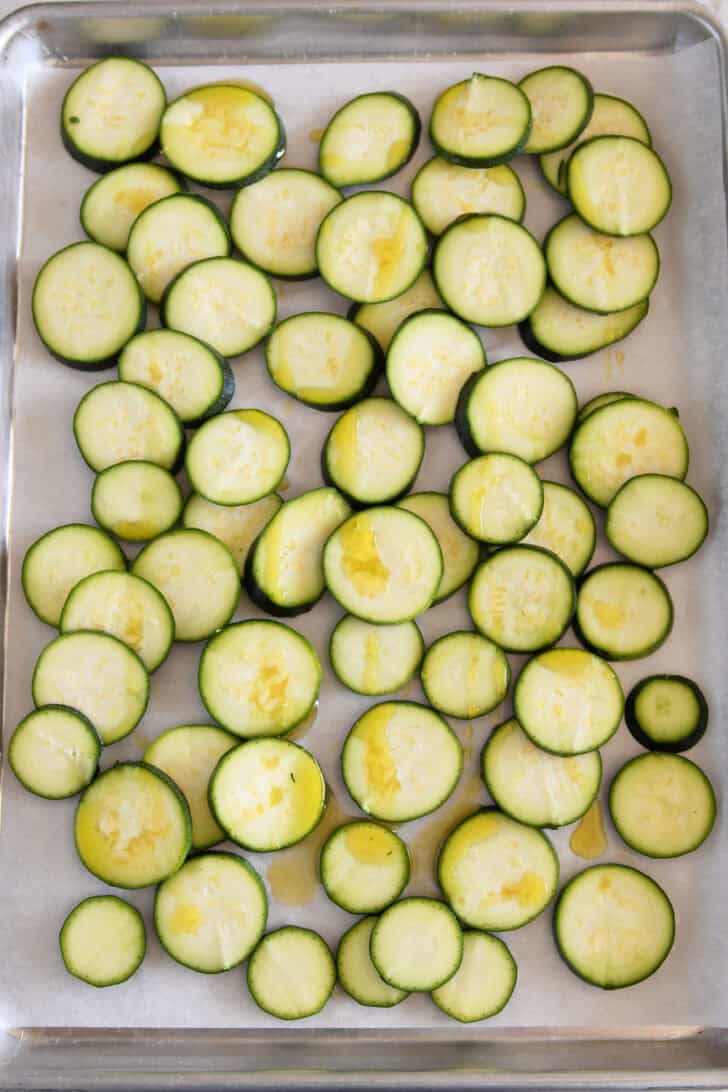 Thinly sliced zucchini rounds on parchment-lined half sheet pan drizzled with olive oil.