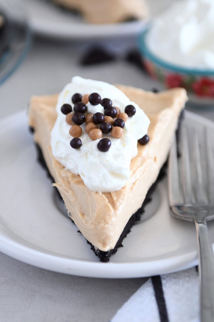 A slice of chocolate cookie crust peanut butter pie on a white plate. The pie is topped with whipped cream and small chocolate balls.