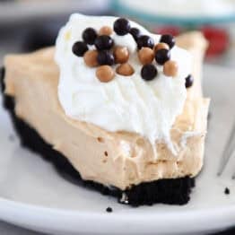 A slice of chocolate cookie crust peanut butter pie with a bite taken out on a white plate. The pie is topped with whipped cream and small chocolate balls.