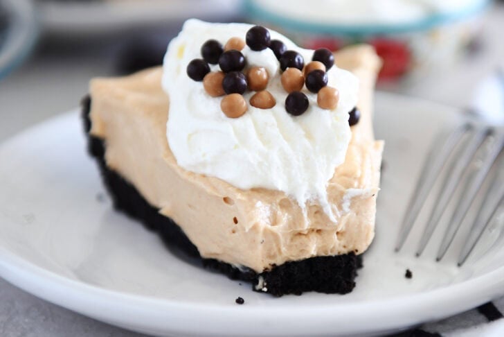 A slice of chocolate cookie crust peanut butter pie with a bite taken out on a white plate. The pie is topped with whipped cream and small chocolate balls.