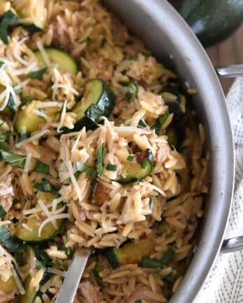 Serving spoon scooping serving of cooked orzo, tuna, zucchini and Parmesan in stainless skillet.