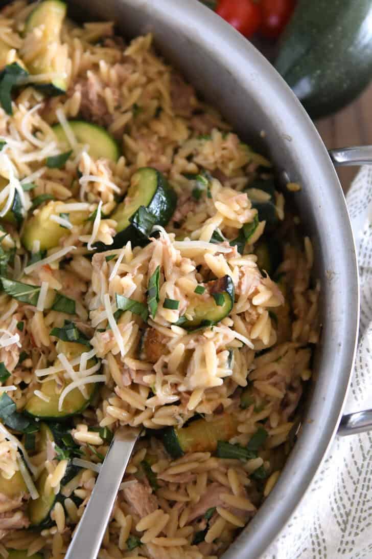 Serving spoon scooping serving of cooked orzo, tuna, zucchini and Parmesan in stainless skillet.
