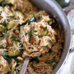 Serving spoon scooping serving of cooked orzo, tuna, zucchini and Parmesan in stainless skillet.