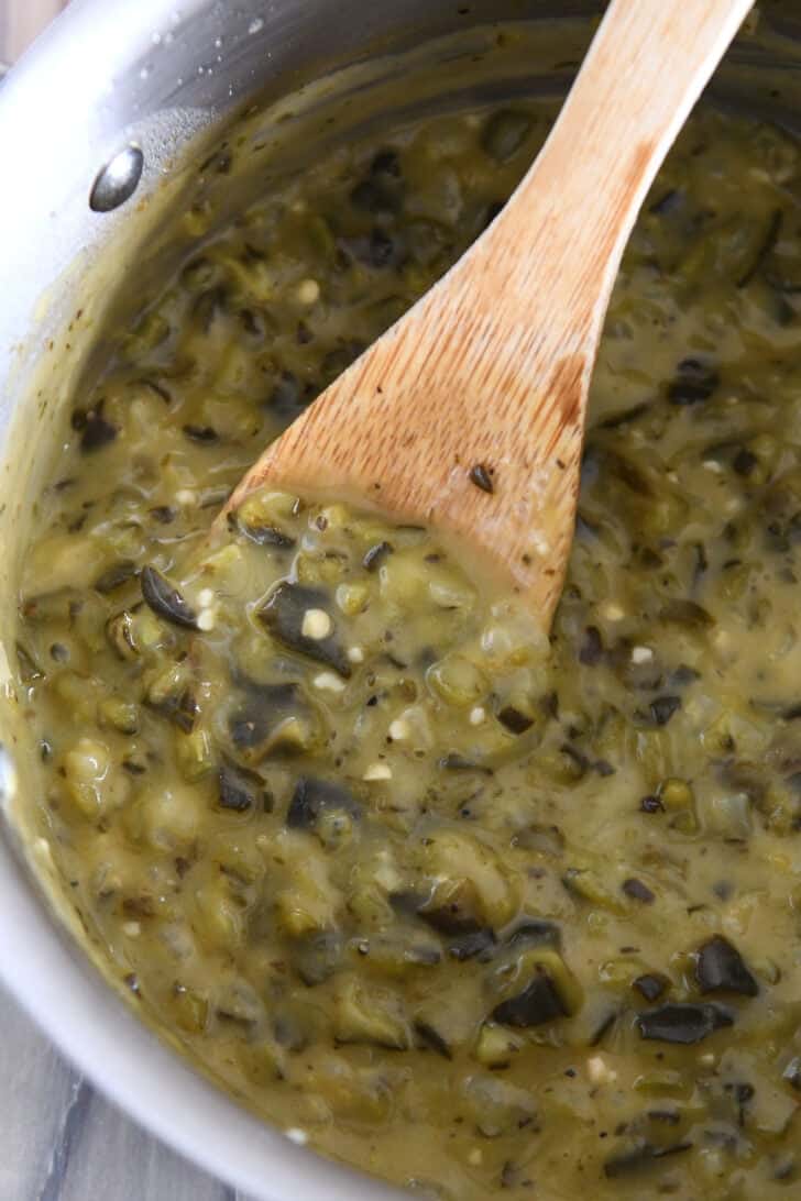 A metal pot with green enchilada sauce cooking and a wooden spoon stirring it.