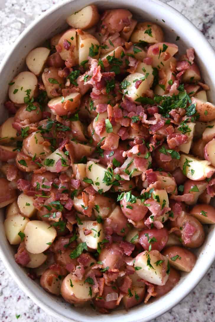 Top down view of a white oval dish with potato chunks, bacon pieces, and chopped parsley.