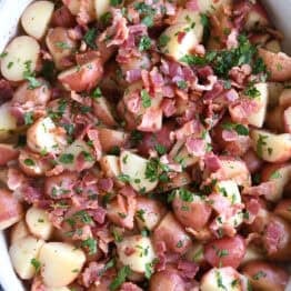 Top down view of a white oval dish with potato chunks, bacon pieces, and chopped parsley.