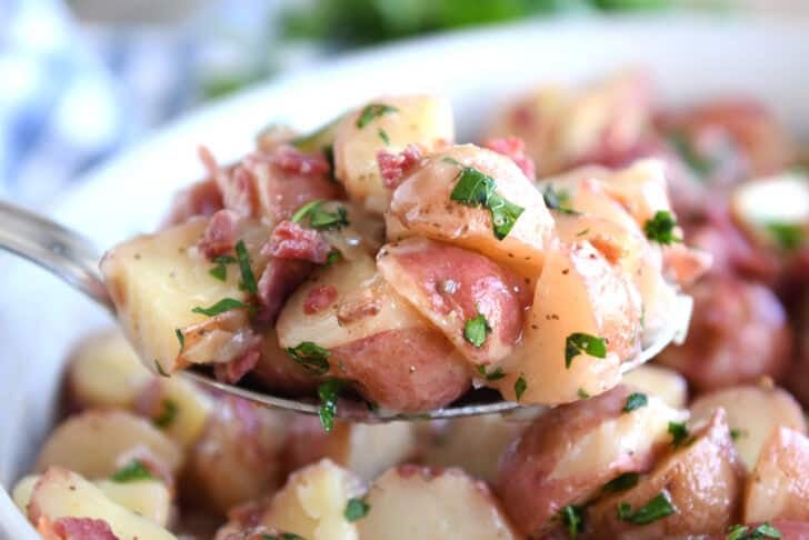 A metal spoon taking a large scoop of german potato salad out of a white dish.