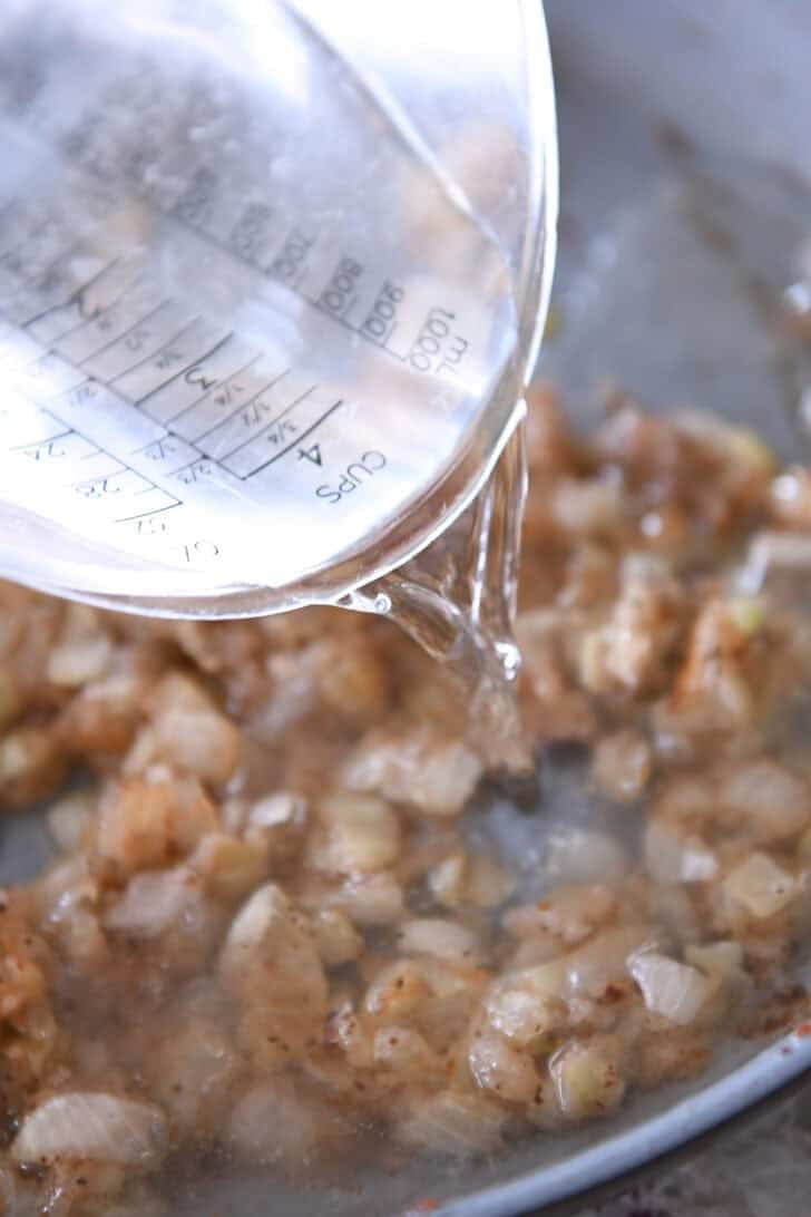 A liquid measuring cup pouring water into a skillet with browned onion pieces.