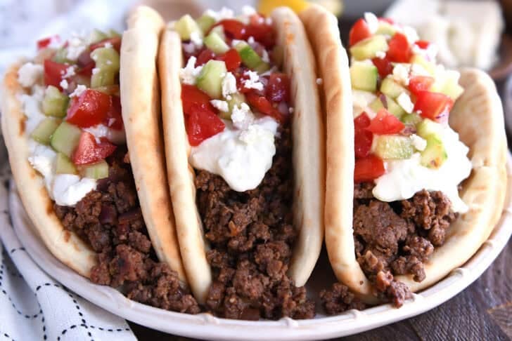 Front view of three stuffed greek flatbread tacos topped with tzatziki sauce, chopped cucumbers, and diced tomatoes.