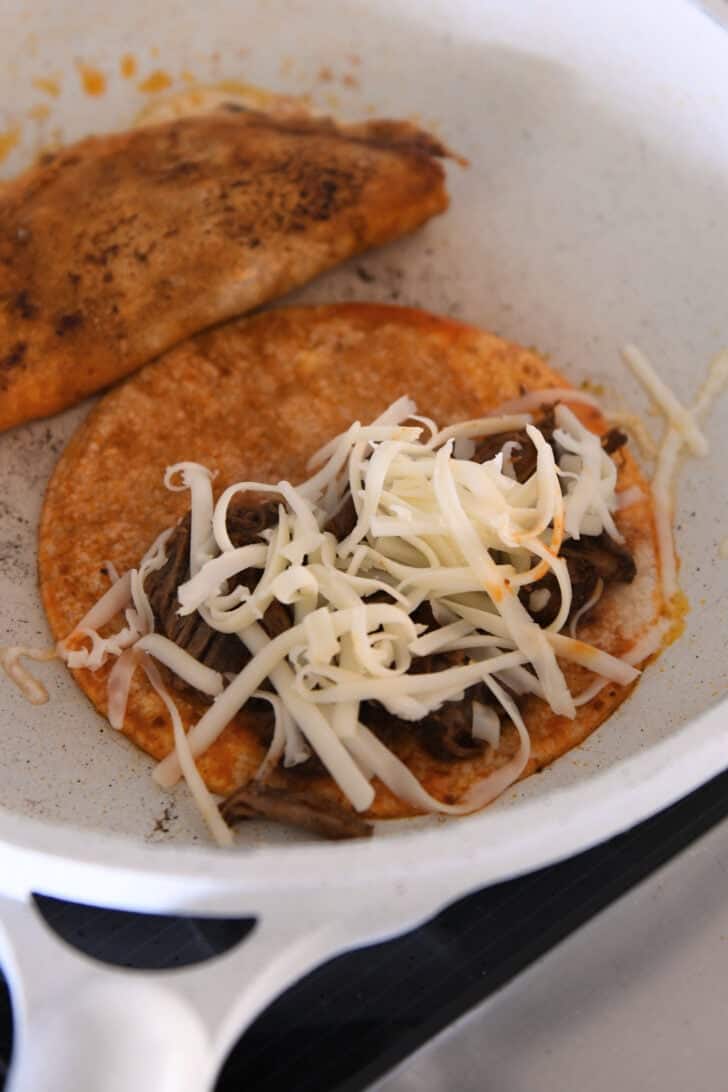 White skillet with two corn tortillas soaked in birria broth topped with shredded beef and shredded cheese.