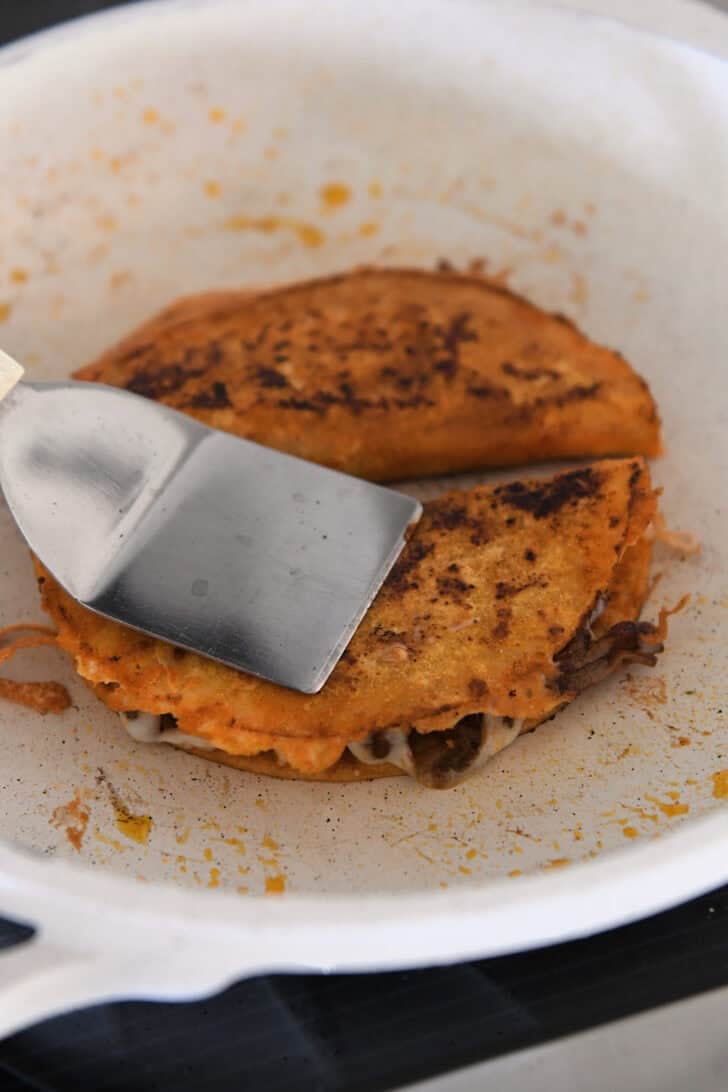Pressing crispy corn tortilla taco in white skillet with metal spatula.
