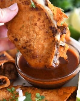 Dipping beef and cheese taco with crispy outer shell in rich birria broth in glass bowl.