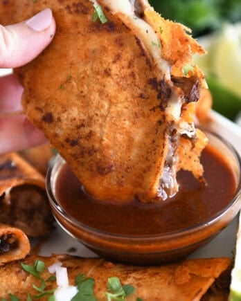 Dipping beef and cheese taco with crispy outer shell in rich birria broth in glass bowl.