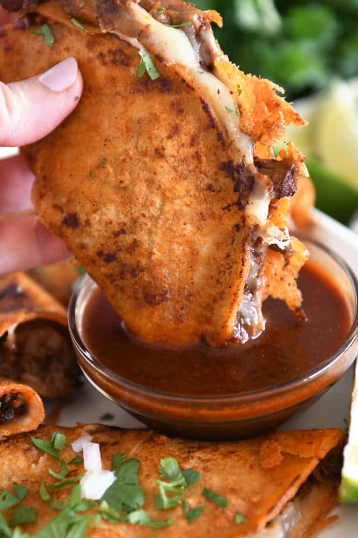 Dipping beef and cheese taco with crispy outer shell in rich birria broth in glass bowl.