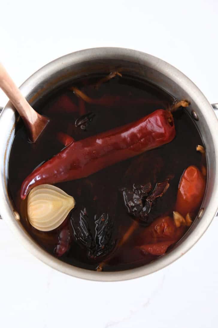 Top down view of pot with simmered broth of guajillo chiles, ancho chiles, arbol chiles, white onion, and garlic.