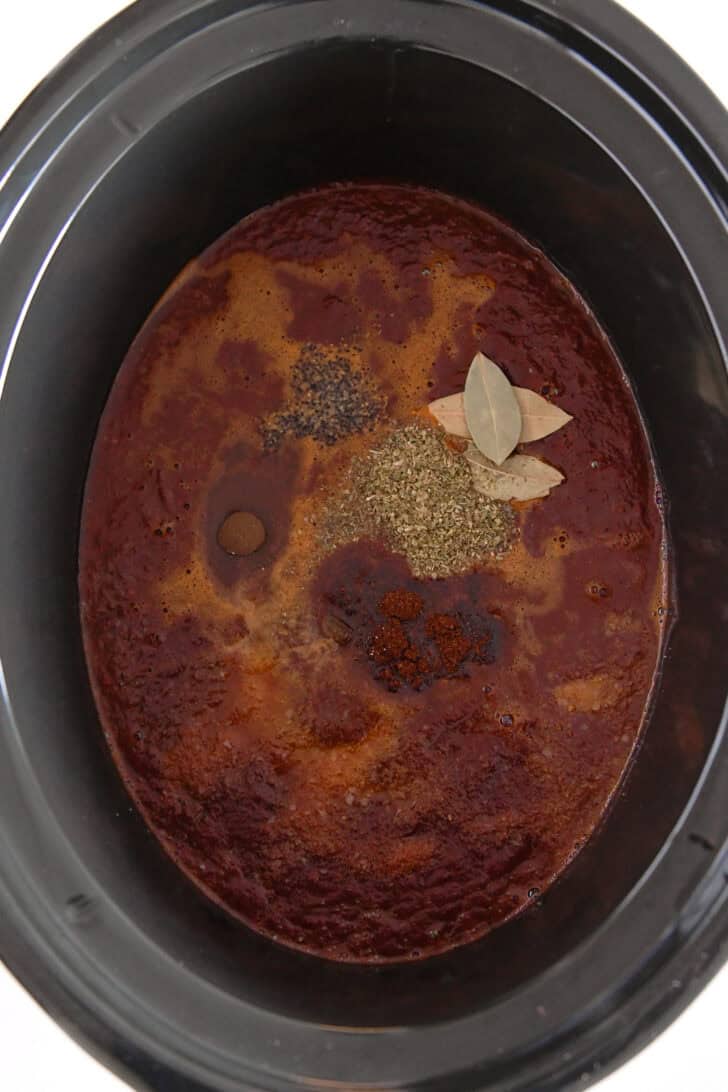 Top down view of black slow cooker insert with dried oregano, bay leaves, chili powder, cumin and blended mixture of Mexican chiles.