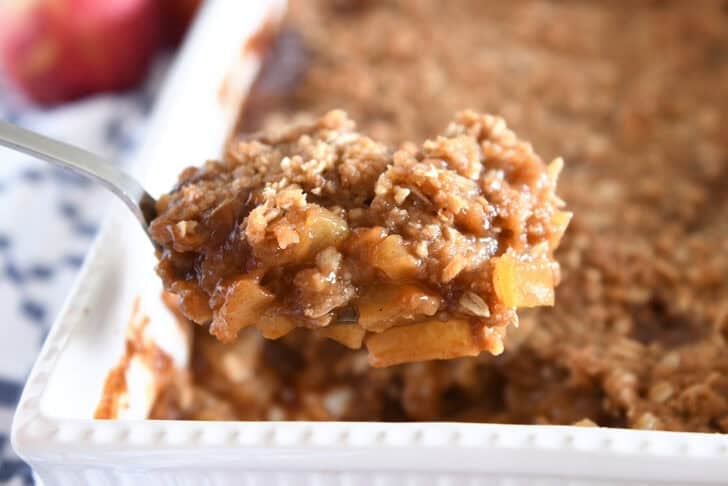 Serving spoon with heaping serving of caramel apples topped with buttery oat crisp in front of pan of caramel apple crisp.