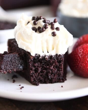 Piece of chocolate olive oil cake topped with whipped cream and crispy chocolate pearls with bite of cake on fork on white plate.