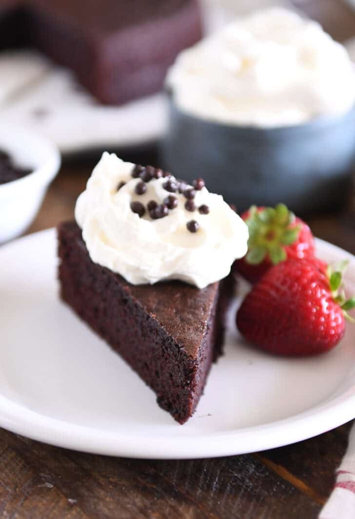 Triangle piece of chocolate olive oil cake on white plate topped with whipped cream and crispy chocolate pearls with strawberries on plate.