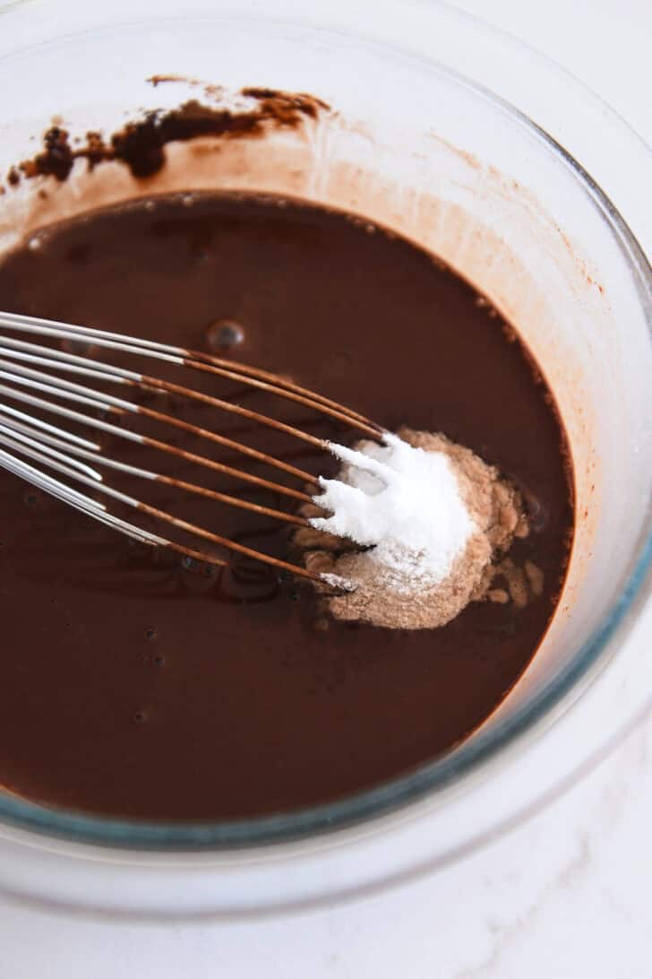 Baking soda and salt added to cocoa powder and water in glass bowl with whisk.