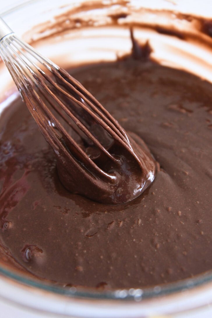 Whisk mixing thick chocolate cake batter in white bowl.