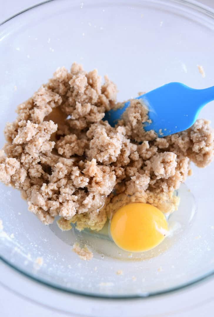 A glass bowl with sugar and butter creamed together, a raw egg, and a blue spatula.