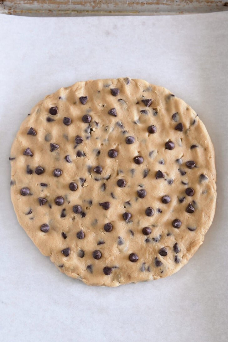 Top down view of a cookie sheet with parchment paper and a large uncooked chocolate chip cookie with extra chocolate chips sprinkled on top in the center.