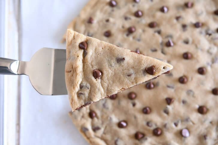 Top down view of a large chocolate chip cookie with a triangle-shaped wedge cut out on a metal spatula.
