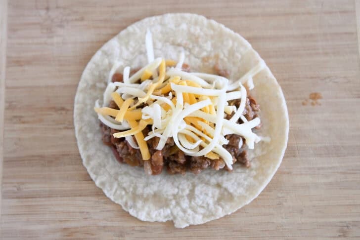 Top down view of a 6-inch flour tortilla topped with ground beef enchilada filling and shredded cheddar and Monterey Jack cheese.