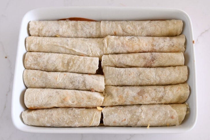 Ten rolled ground beef enchiladas in flour tortillas in white 9X13-inch dish.
