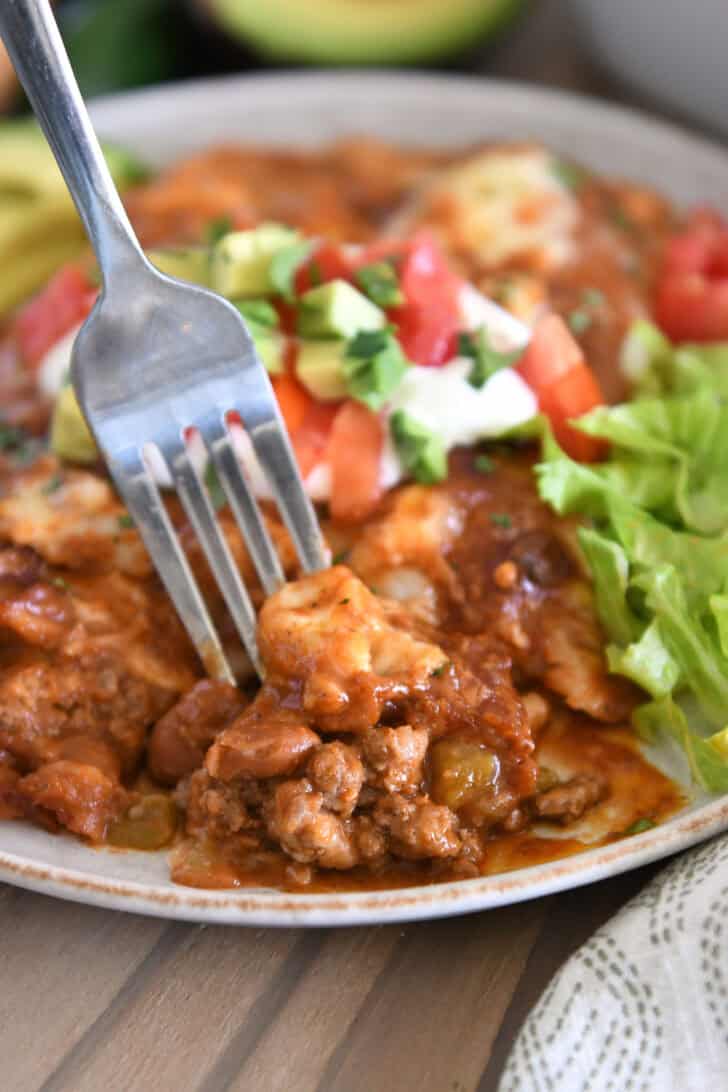 Fork scooping bite of ground beef enchiladas with red sauce on gray plate with lettuce, sour cream, avocados and cilantro.