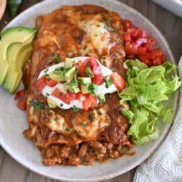 Top down view of two enchiladas on gray plate topped with red enchilada sauce, cheese, cilantro, sour cream, tomatoes, and avocados.