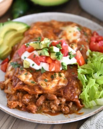 Top down view of two enchiladas on gray plate topped with red enchilada sauce, cheese, cilantro, sour cream, tomatoes, and avocados.