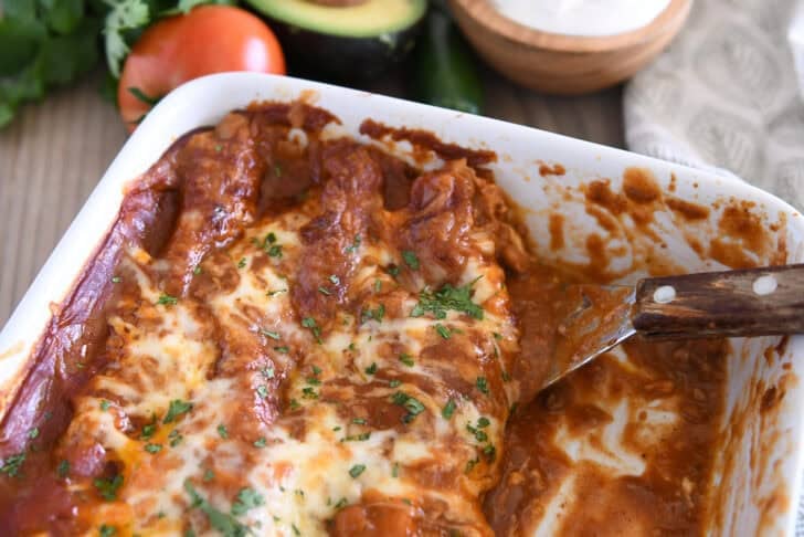 Square metal spatula scooping red sauce ground beef enchiladas from white 9X13-inch pan.