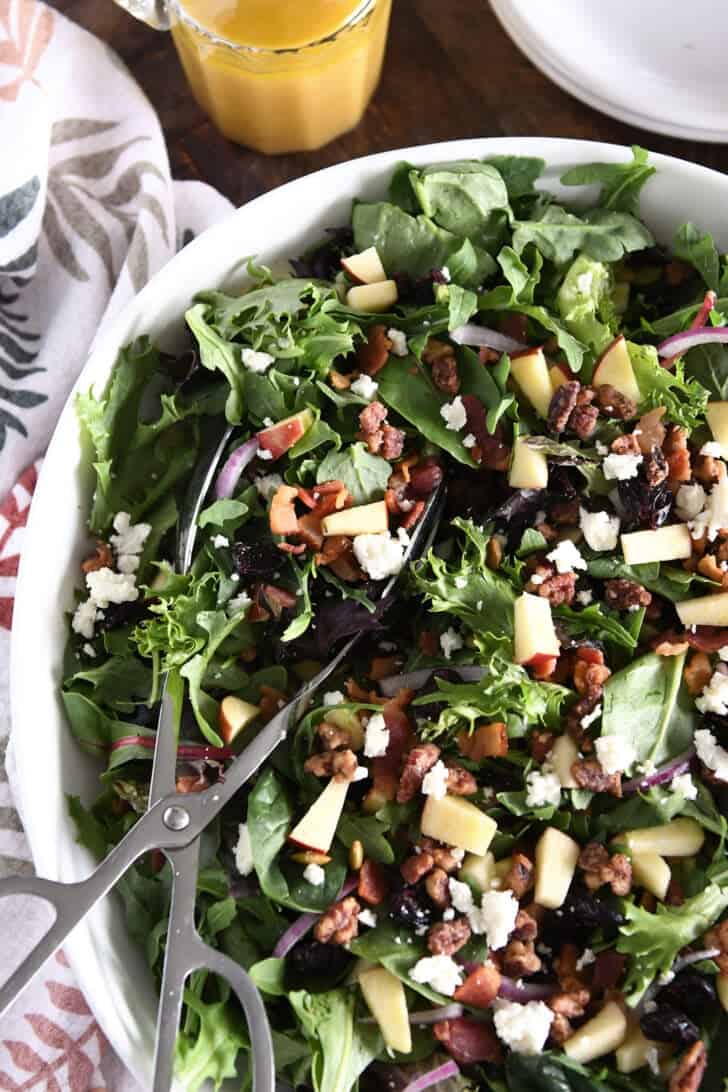 White bowl with salad tongs and mixed salad of baby greens, chopped apples, feta cheese, cooked bacon, craisins, and pumpkin seeds.