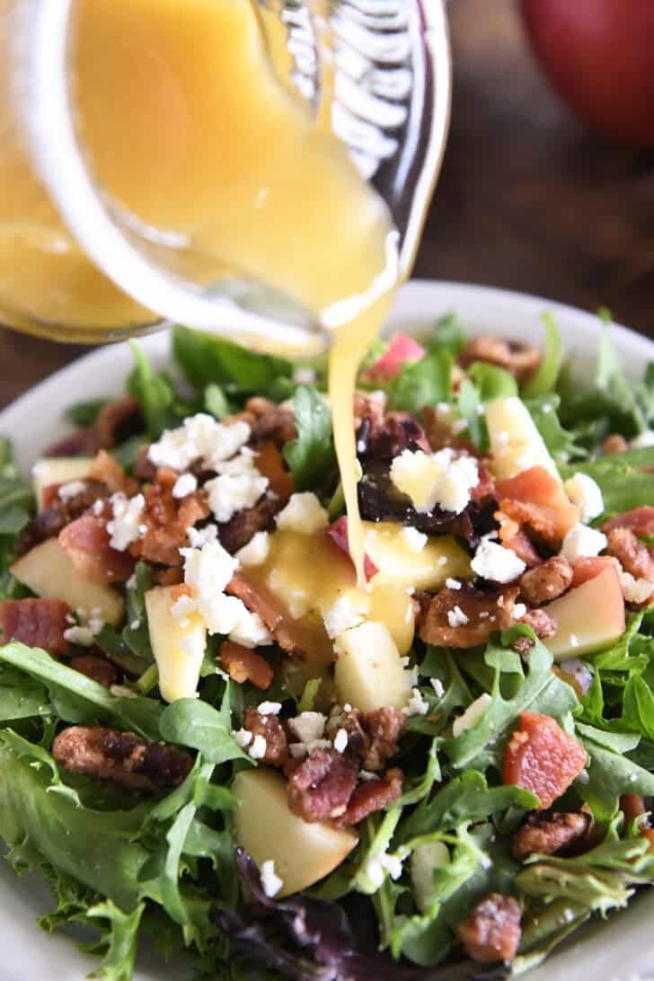 Pouring apple cider vinaigrette over green salad with chopped apples, feta cheese, pecans and bacon.