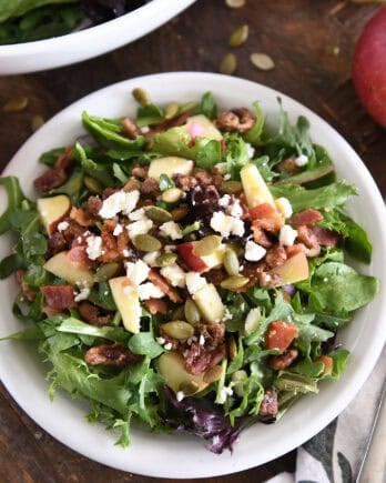 Top down view of green salad on white plate with roasted pumpkin seeds, chopped apples, candied pecans, feta cheese and red onions.