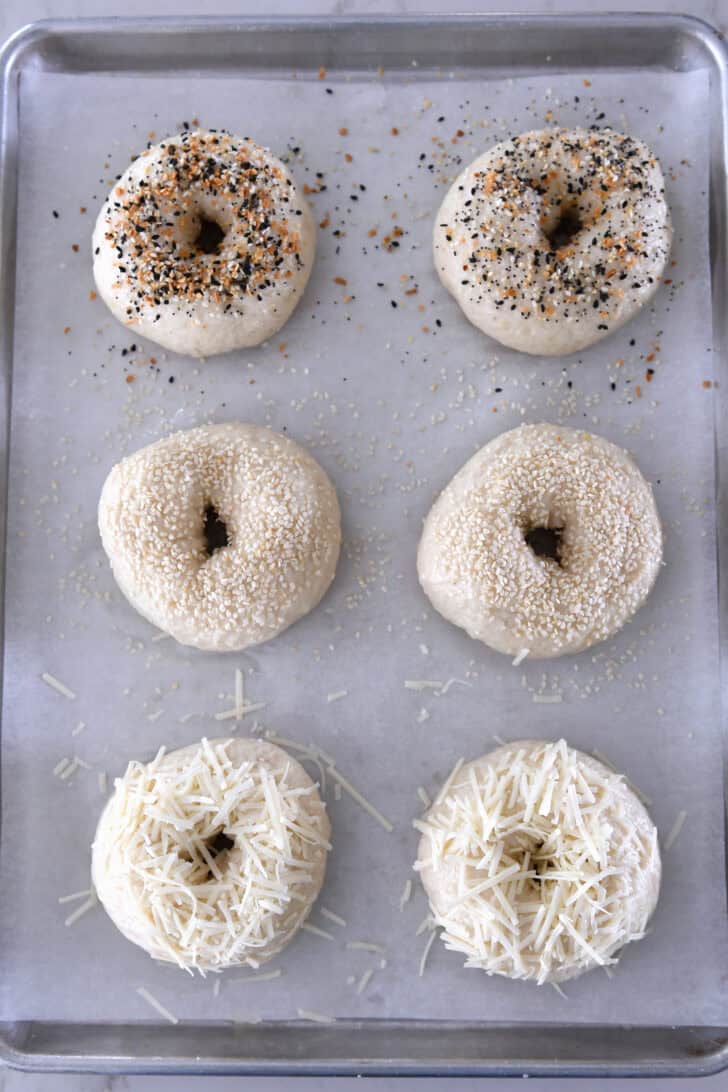 Top down view of six unbaked bagels. The top two have everything bagel seasoning, the middle two have sesame seeds and the bottom two have shredded Parmesan cheese.
