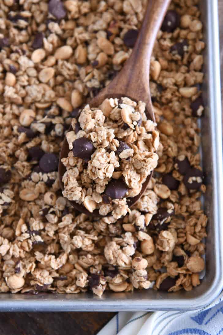 Wooden spoon filled with peanut butter granola with roasted peanuts and dark chocolate chunks resting on sheet pan of granola.