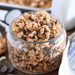 Shallow glass jar filled with peanut butter granola with roasted peanuts and dark chocolate chunks with spoonful of peanut butter to the side.