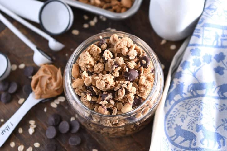 Top down view of shallow glass jar filled with peanut butter granola with roasted peanuts and dark chocolate chunks with spoonful of peanut butter to the side.