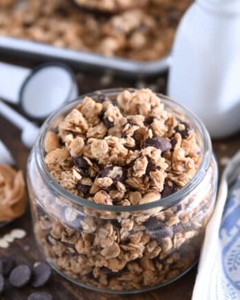 Shallow glass jar filled with peanut butter granola with roasted peanuts and dark chocolate chunks with spoonful of peanut butter to the side.