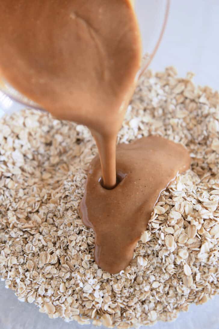 Pouring peanut butter syrup over dry oats in glass bowl.