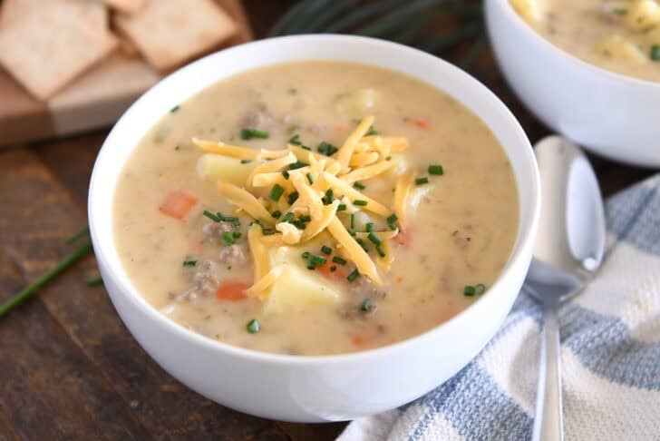 White bowl with creamy cheesy soup with cooked potatoes, carrots and ground beef topped with shredded cheddar cheese and chopped chives.