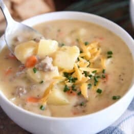 White bowl with spoon scooping ingredients in creamy cheesy soup with cooked potatoes, ground beef, and carrots and topped with shredded cheddar cheese and chopped chives.