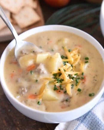 White bowl with spoon scooping ingredients in creamy cheesy soup with cooked potatoes, ground beef, and carrots and topped with shredded cheddar cheese and chopped chives.