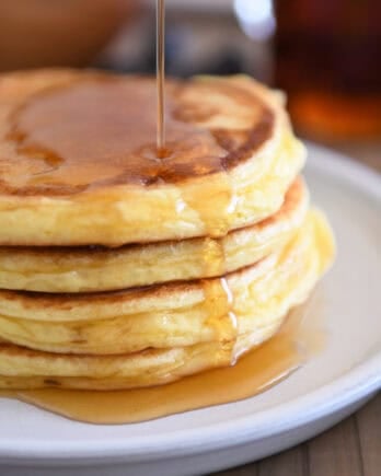 Drizzling pure maple syrup over flour stacked cottage cheese pancakes on gray plate.