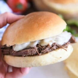 Slow cooker beef on crusty rolls with provolone cheese and one bite taken out; hand holding sandwich.